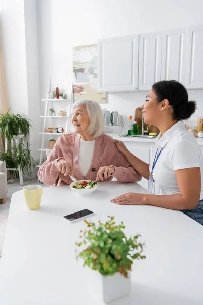 Happy senior woman with grey hair having lunch next to cheerful multiracial social worker — Stock Photo
