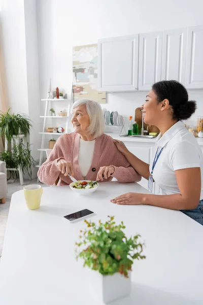 Mulher sênior feliz com cabelos grisalhos almoçando ao lado de assistente social multirracial alegre — Fotografia de Stock