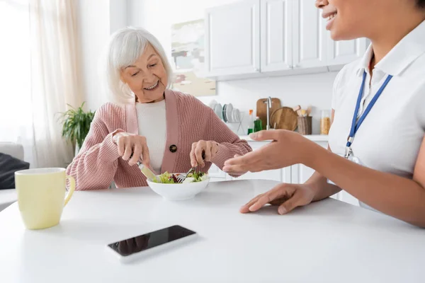 Happy senior woman with grey hair having lunch next to cheerful multiracial caregiver — Stock Photo