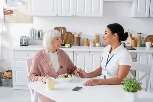 Femme à la retraite avec les cheveux gris tenant la main avec soignant multiracial joyeux à côté du déjeuner sur la table — Photo de stock