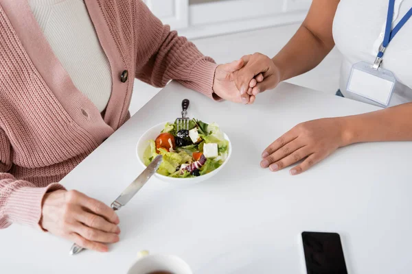 Vue du dessus de la femme âgée tenant la main avec l'aidant multiracial à côté du déjeuner sur la table — Photo de stock