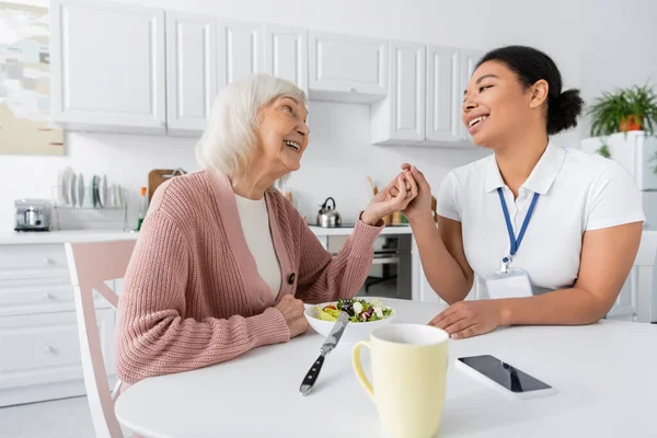 Happy retired woman with grey hair holding hands with cheerful multiracial social worker next to lunch on table — Stock Photo