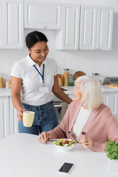 Happy multiracial social worker holding cup of tea near senior woman with grey hair having lunch in kitchen — Stock Photo