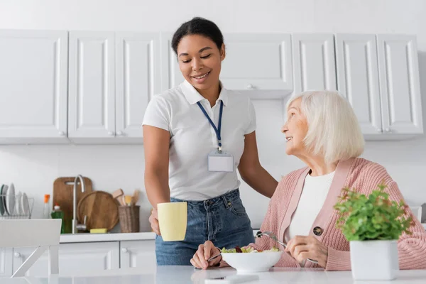 Щасливий багаторасовий соціальний працівник тримає чашку чаю біля пенсіонерів з сірим волоссям обідаючи на кухні — стокове фото