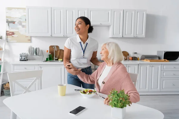 Travailleur social multiracial joyeux tenant la main d'une femme retraitée reconnaissante dans la cuisine — Photo de stock