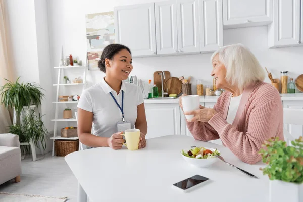 Happy multiracial social worker having tea with senior woman in kitchen — Stock Photo