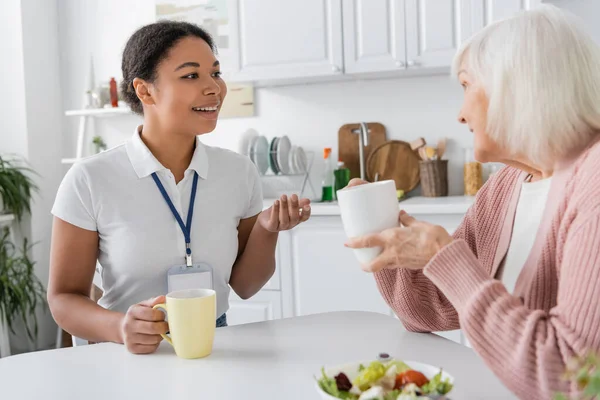 Fröhliche multiethnische Sozialarbeiterin im Gespräch mit einer Seniorin bei einer Tasse Tee — Stockfoto
