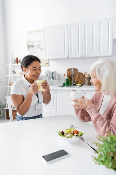Happy multiracial social worker having tea with positive senior woman in kitchen — Stock Photo