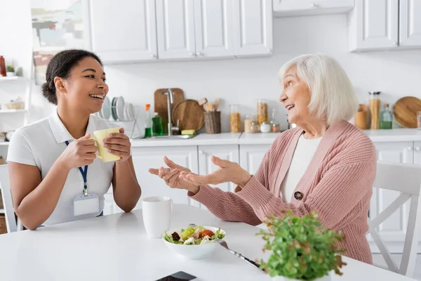 Happy senior woman having tea during conversation with multiracial social worker in kitchen — Stock Photo