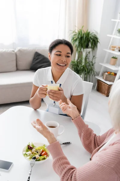 Happy senior woman having conversation with multiracial social worker in kitchen — Stock Photo