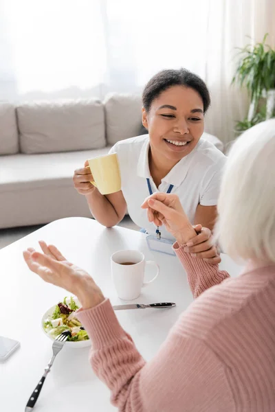 Happy multiracial social worker having tea during conversation with senior woman in kitchen — Stock Photo