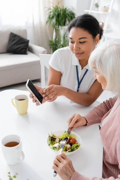 Happy multiracial social worker pointing at smartphone near senior woman having lunch in kitchen — Stock Photo