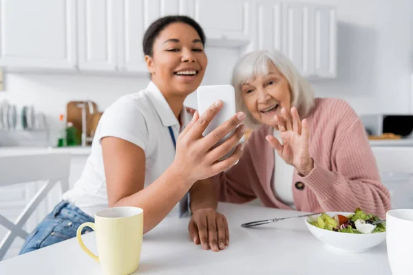 Heureux travailleur social multiracial tenant smartphone tandis que la femme âgée agitant la main lors d'un appel vidéo — Photo de stock