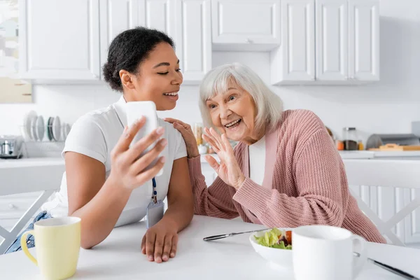 Travailleur social multiracial joyeux tenant smartphone tandis que la femme âgée agitant la main lors d'un appel vidéo — Photo de stock
