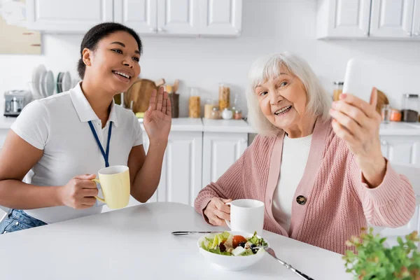 Feliz anciana sosteniendo teléfono inteligente mientras alegre trabajador social multirracial saludando la mano durante la videollamada - foto de stock