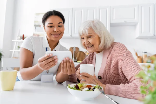 Fröhlicher multiethnischer Sozialarbeiter zeigt Rentnerin beim Mittagessen in Küche Smartphone — Stockfoto