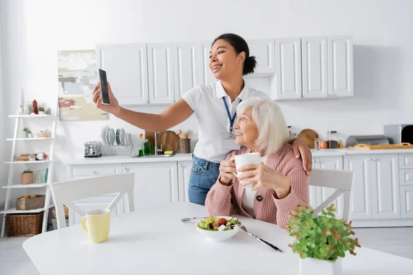 Fröhliche multiethnische Sozialarbeiterin macht Selfie mit Rentnerin während des Mittagessens in der Küche — Stockfoto