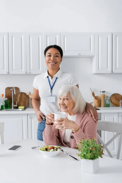 Heureux multiracial travailleur social souriant avec la femme retraitée pendant le déjeuner dans la cuisine — Photo de stock