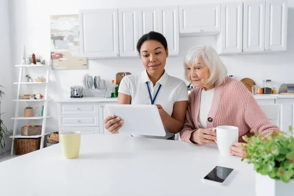 Felice assistente sociale multirazziale in possesso di tablet digitale vicino alla donna anziana in cucina — Foto stock