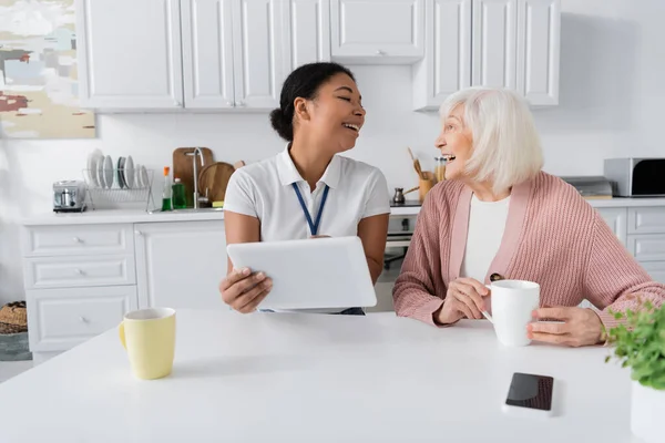 Веселий багаторасовий соціальний працівник тримає цифровий планшет біля старшої жінки на кухні — стокове фото