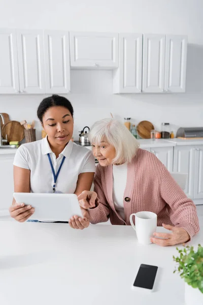 Щасливий багаторасовий соціальний працівник, що показує цифровий планшет старшій жінці на кухні — стокове фото