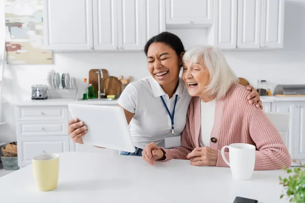Happy multiracial social worker holding digital tablet and laughing with senior woman in kitchen — Stock Photo