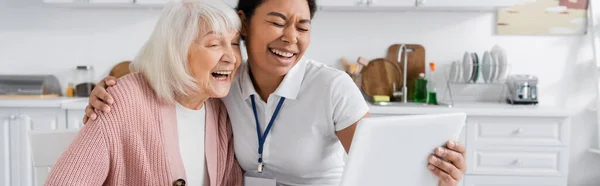 Feliz trabajador social multirracial sosteniendo tableta digital y riendo con la mujer mayor en la cocina, pancarta — Stock Photo