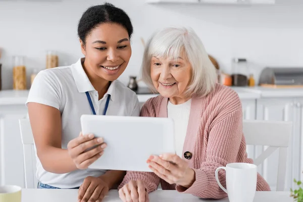 Gioioso assistente sociale multirazziale in possesso di tablet digitale vicino alla donna anziana in cucina — Foto stock