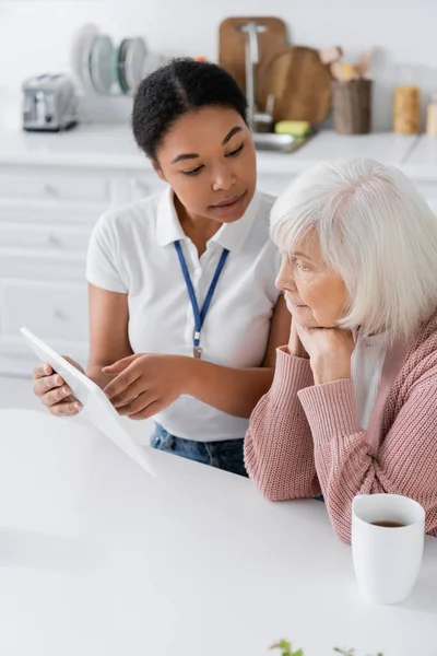 Positive multiracial social worker holding digital tablet near senior woman in kitchen — Stock Photo