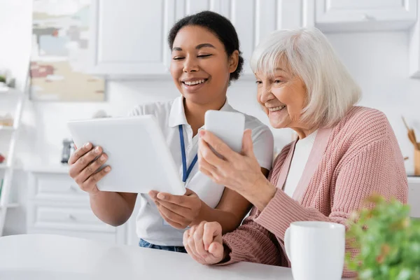 Feliz trabajador social multirracial sosteniendo tableta digital cerca de la mujer mayor con teléfono inteligente en la cocina - foto de stock