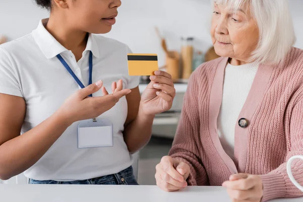 Travailleur social multiracial détenant une carte de crédit près d'une femme âgée à la maison — Photo de stock