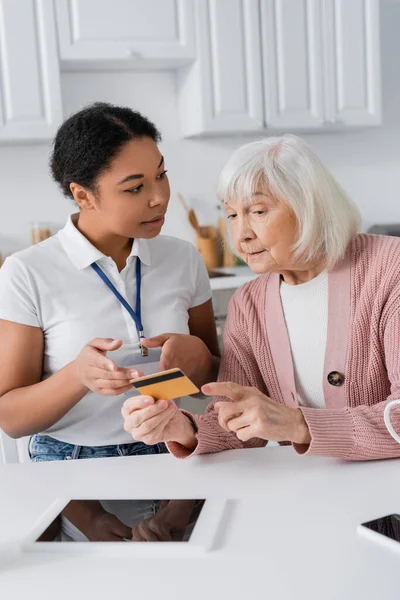 Brünette Sozialarbeiterin mit Kreditkarte in der Nähe von Seniorin zu Hause — Stockfoto