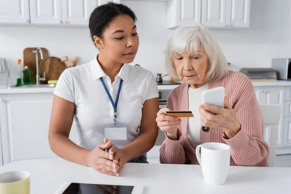Morena multirracial trabajador social mirando a la mujer mayor con tarjeta de crédito y teléfono inteligente haciendo compras en línea - foto de stock