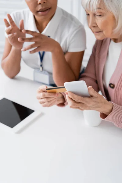 Trabajador social multirracial explicando a la mujer mayor con tarjeta de crédito y teléfono inteligente cómo hacer compras en línea - foto de stock