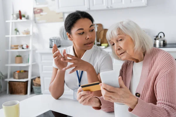 Multiracial social worker explaining retired woman with credit card and smartphone how to do online shopping — Stock Photo
