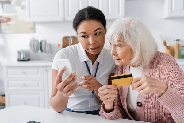 Morena multirracial trabajador social sosteniendo teléfono inteligente cerca de la mujer mayor con tarjeta de crédito - foto de stock