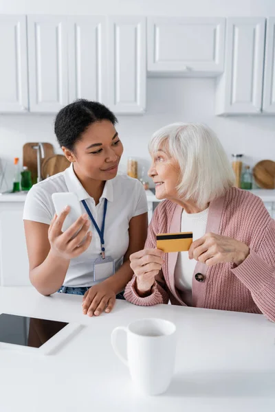 Morena multirracial trabajador social sosteniendo teléfono inteligente cerca feliz mujer mayor con tarjeta de crédito - foto de stock