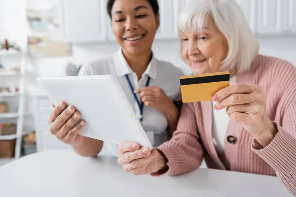 Брюнетка багаторасовий соціальний працівник тримає цифровий планшет біля щасливої старшої жінки з кредитною карткою — стокове фото