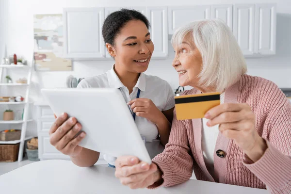Allegro assistente sociale multirazziale in possesso di tablet digitale vicino felice donna anziana con carta di credito — Foto stock
