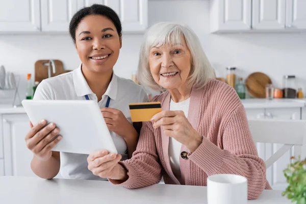 Жизнерадостный мультирасовый социальный работник, держащий цифровой планшет рядом с положительной пожилой женщиной с кредитной картой — стоковое фото