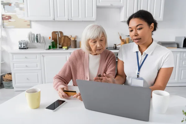 Trabajador social multirracial utilizando el ordenador portátil al explicar a la mujer mayor con tarjeta de crédito cómo hacer compras en línea - foto de stock