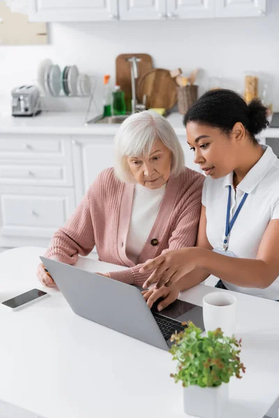 Multiracial social worker explaining how to use laptop to senior woman with credit card — Stock Photo