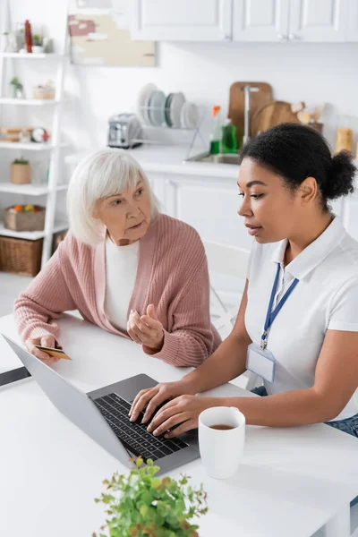 Multiracial social worker using laptop near senior woman with credit card — Stock Photo