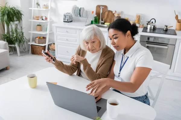 Assistente social multirracial usando laptop perto de mulher aposentada com smartphone — Fotografia de Stock