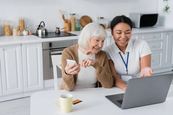 Trabajador social multirracial feliz usando el ordenador portátil cerca de la mujer jubilada con teléfono inteligente - foto de stock