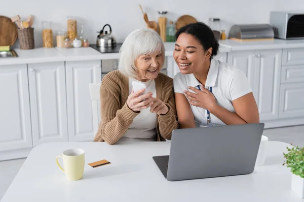 Fröhliche multiethnische Sozialarbeiterin blickt auf Smartphone in den Händen einer Rentnerin in der Nähe von Laptop — Stockfoto