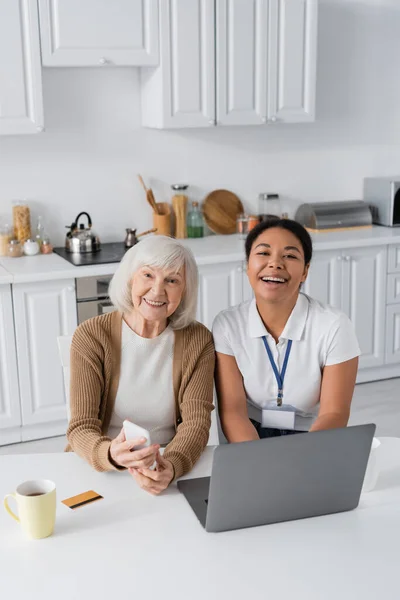 Travailleur social multiracial joyeux et femme âgée tenant smartphone près d'un ordinateur portable sur la table — Photo de stock