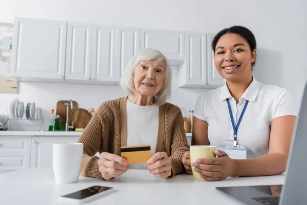 Fröhliche multiethnische Sozialarbeiterin hält Tasse neben Seniorin mit Kreditkarte und Gadgets — Stockfoto