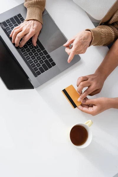 Вид женщины на пенсии, печатающей на клавиатуре ноутбука рядом с мультирасовым социальным работником с помощью кредитной карты — стоковое фото