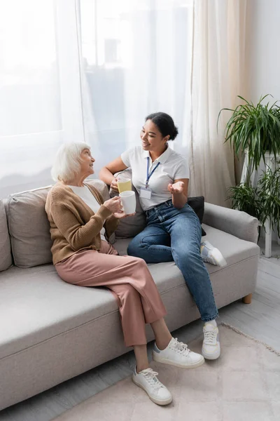 Fröhliche multirassische Sozialarbeiterin plaudert mit Seniorin beim Tee im Wohnzimmer — Stockfoto